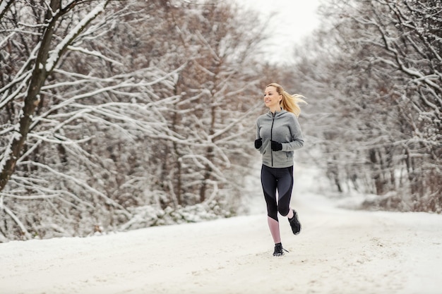 In voller Länge von einer glücklichen Frau mittleren Alters, die im Winter auf verschneitem Weg in der Natur läuft. Winterfitness, gesunde Gewohnheiten