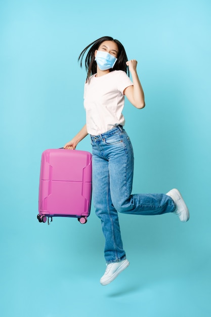 In voller Länge von glücklichen asiatischen Touristen, tanzen und springen mit Koffer in medizinischer Gesichtsmaske, aufgeregt über Urlaub, auf blauem Hintergrund stehend.