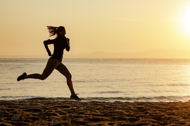 In voller Länge von motivierter athletischer Frau, die bei Sonnenuntergang am Strand läuft Kopieren Sie Platz