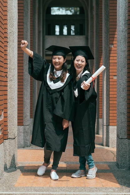 in voller länge zwei schöne lächelnde junge studentin mit diplom, die mit einem freund mit in der faust in der luft erhobenen armen steht. college-frau am abschlusstag in kleidern und kappe gesicht kamera lächelnd feiern