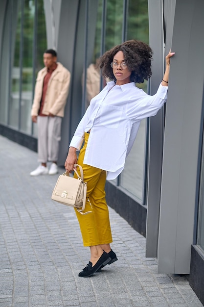 Inacessível. Jovem morena de cabelos encaracolados com óculos com roupa formal elegante em pé posando perto do prédio da cidade e homem indeciso atrás