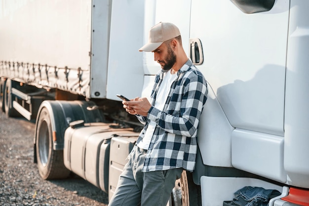 Inclinando-se ao lado do veículo e usando o telefone Jovem caminhoneiro está de dia