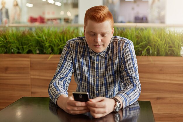 Un inconformista elegante con el pelo rojo de moda y pecas vestidas con camisa a cuadros sentado en el café.