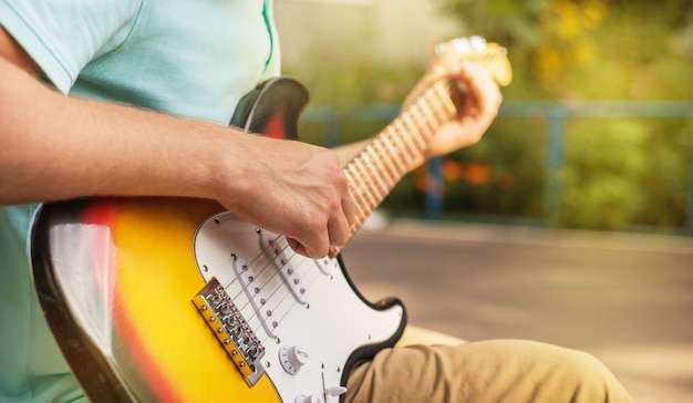 Foto inconformista feliz del hombre joven que se sienta tocando la guitarra, cierre para arriba, luz del sol del verano, al aire libre