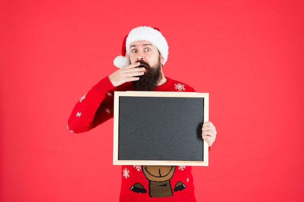 Increíble. Anuncio de invierno. Evento de invierno. Vacaciones de invierno. Sombrero de Papá Noel con publicidad. Pizarra para información. Hombre mantenga espacio de copia de pizarra en blanco. Guy santa claus fondo rojo.