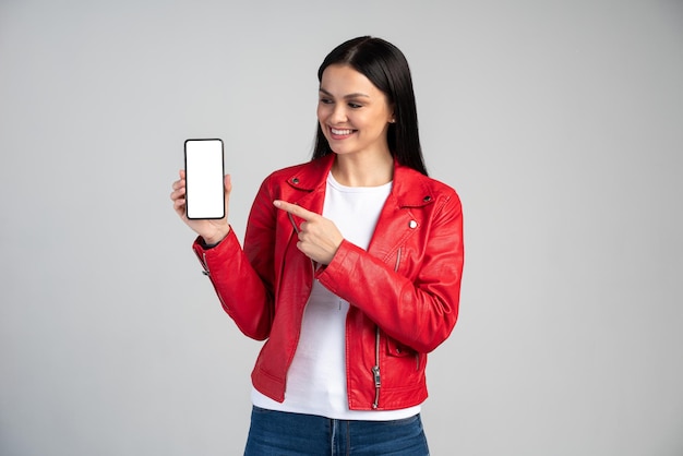 Increíble aplicación móvil. Mujer linda emocionada sorprendida apuntando al teléfono celular y mirándolo con cara sonriente, emocionada por teléfono, servicio en línea. Tiro de estudio interior fondo gris