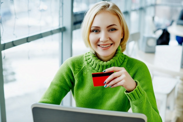 Increíble chica con cabello claro con suéter verde sentado en la cafetería con computadora portátil y tarjeta de crédito, retrato, concepto independiente, compras en línea.