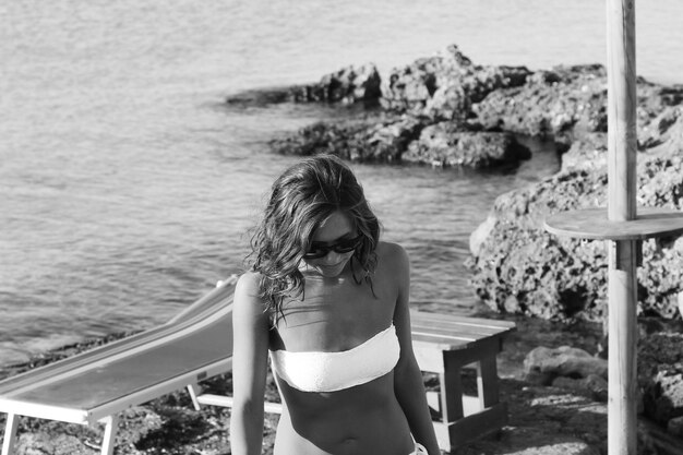 Increíble chica con gafas de sol en un traje de baño blanco caminando por la playa rocosa Apulia Salento Italia