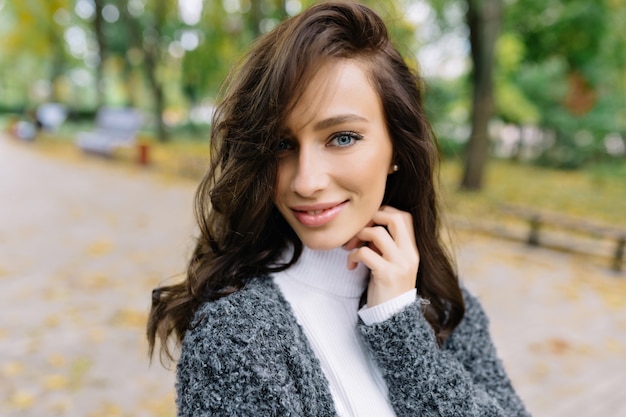 Increíble chica guapa alegre con cabello corto morena sonriendo a la cámara en el fondo de la ciudad. La mujer joven alegre de moda ha tejido suéter gris, emociones verdaderas positivas