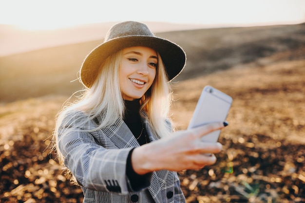 Increíble mujer caucásica rubia con sombrero haciendo un selfie con smartphone contra una hermosa puesta de sol mientras viaja.