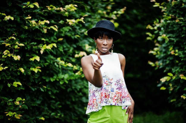 Increíble mujer modelo afroamericana con pantalones verdes y sombrero negro posando con diferentes emociones en el parque muestra un pulgar hacia arriba