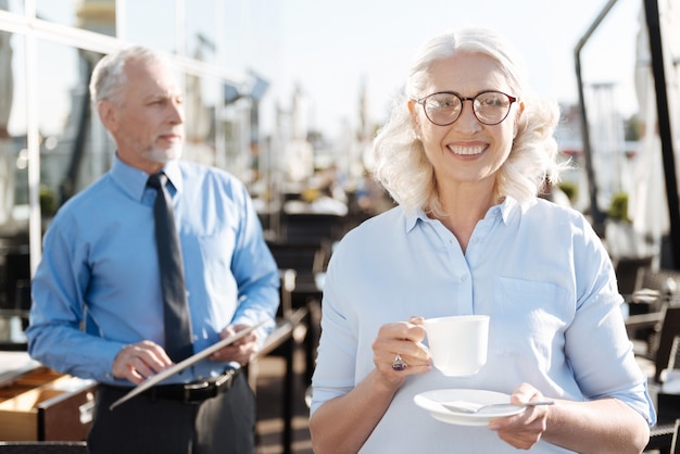 Increíble mujer posando con taza en las manos mientras usa gafas elegantes y siente felicidad