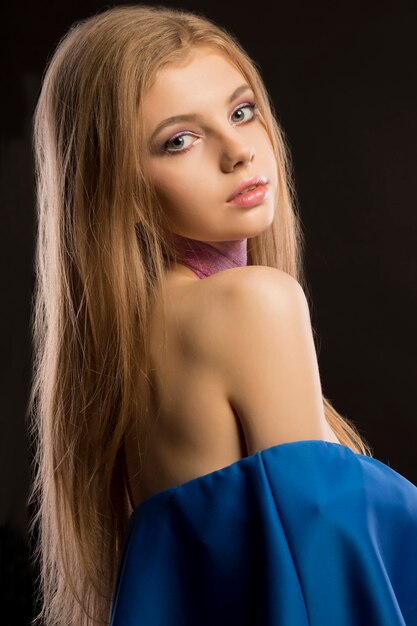 Increíble mujer rubia con cabello largo y exuberante y vestido azul con hombros desnudos posando en el estudio