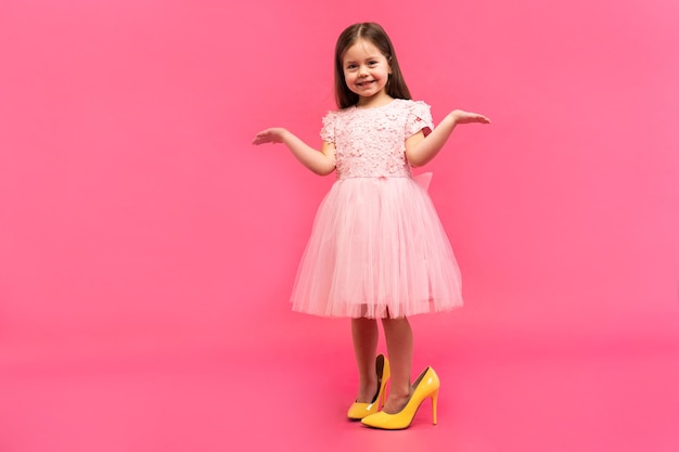 Increíble niña feliz emocionada en vestido con zapatos de mamá expresando a cámara aislada sobre fondo rosa. Emociones brillantes.