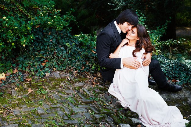 Increíble novia con el pelo largo y rizado y el novio sentado cerca uno del otro en el fondo de hojas verdes, foto de boda, hermosa pareja, día de la boda, retrato de cerca.