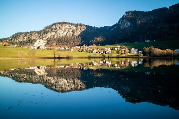 Increíble paisaje tranquilo con área de campo, casas y colinas cerca del lago y reflejo en agua azul limpia sobre un fondo de cielo azul claro, Austria.