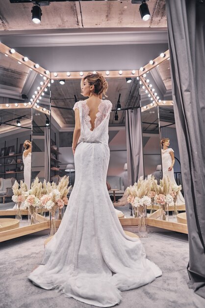 Increíblemente hermoso. Vista trasera de longitud completa de atractivas mujeres jóvenes con un vestido de novia increíble mientras está de pie frente al espejo en la tienda nupcial