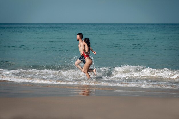 Incríveis recém-casados na praia exótica correndo alegremente nas águas rasas, espirra e risos ao redor.