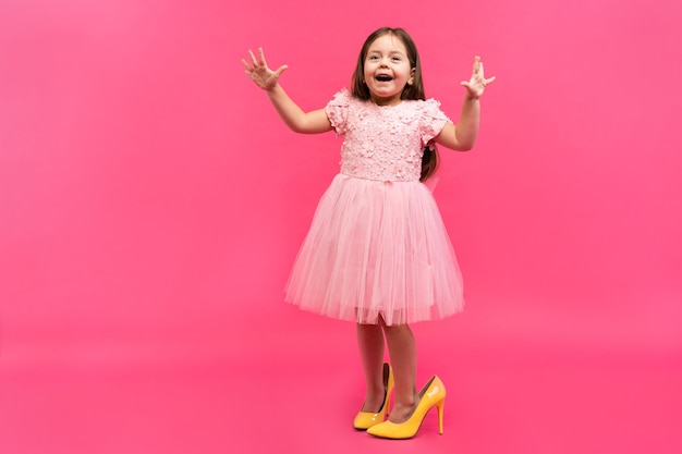 Incrível garota feliz animada no vestido com os sapatos da mamãe, expressando-se para a câmera isolada no fundo rosa. Emoções brilhantes.