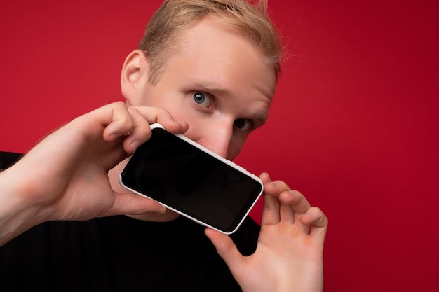 Incrível homem loiro adulto bonito vestindo camiseta preta em pé isolado em um fundo vermelho com espaço de cópia, segurando o smartphone, mostrando o telefone na mão com tela vazia para maquete, olhando para a câmera.
