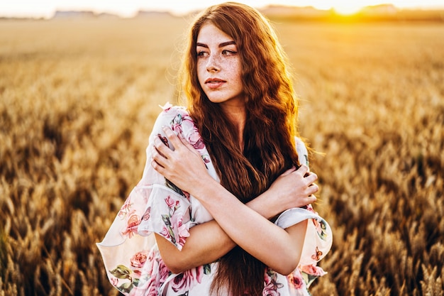 Incrível jovem com rosto longo cabelo encaracolado e sardas. Mulher de vestido posando no campo de trigo ao pôr do sol. Fechar retrato