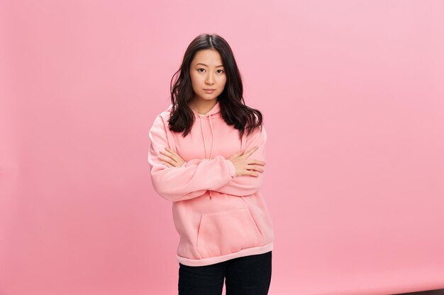Incrível linda e alegre estudante asiática feliz jovem em moletom com capuz rosa dobra as mãos posando isoladas sobre fundo de estúdio rosa boa oferta conceito de nova coleção de moda