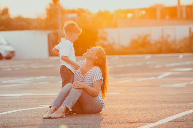 Incrível retrato de verão da jovem mãe feliz e seu adorável filho olhando um ao outro sentado no asfalto no estacionamento vazio em uma luz dourada do sol.