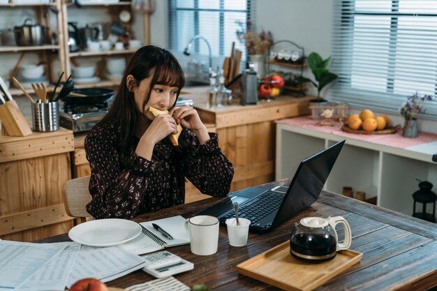 La independiente asiática está programando su nuevo programa de vacaciones entrantes y desayuna al mismo tiempo frente a la cocina. trabajo desde el concepto de hogar.