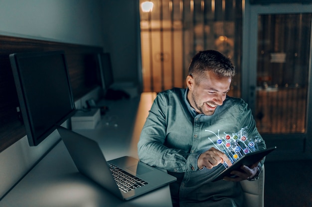 Independiente barbudo caucásico guapo con sonrisa toothy sentado en la oficina a altas horas de la noche y usando tableta para navegar por internet. concepto de redes sociales.