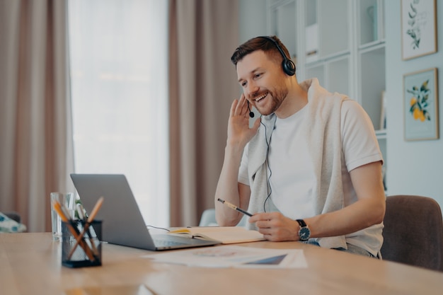 Independiente masculino positivo o poses de estudiante en la oficina en casa usa auriculares participa en el seminario web de educación hace que las notas se vean felices en la pantalla del portátil hace que la videollamada se vista de manera informal comunicación web