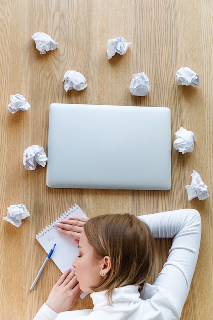 Independiente mujer cansada durmiendo y descansando su cabeza sobre el cuaderno sobre la mesa, escribiendo en el bloc de notas, bolas de papel arrugadas y portátil alrededor, vista superior.