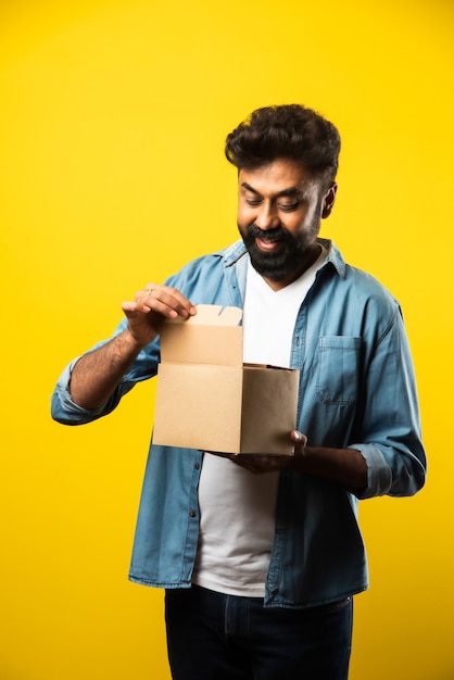 Indio guapo joven barbudo abriendo la caja con el paquete mientras está de pie en amarillo