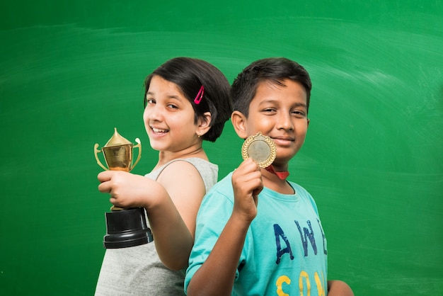 Indische asiatische Schulkinder, die Goldtrophäenbecher über grünem Tafelhintergrund halten