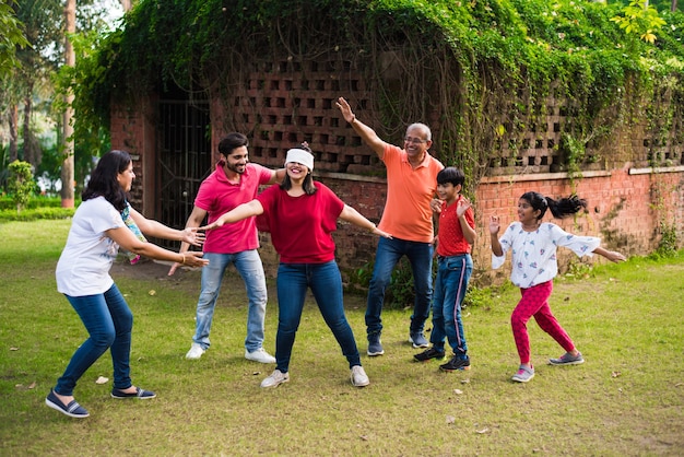 Indische Familie mit verbundenen Augen oder andhali Koshimbir-Spiel im Park oder Garten, asiatische Familie mit mehreren Generationen, die Spaßspiele im Freien spielt