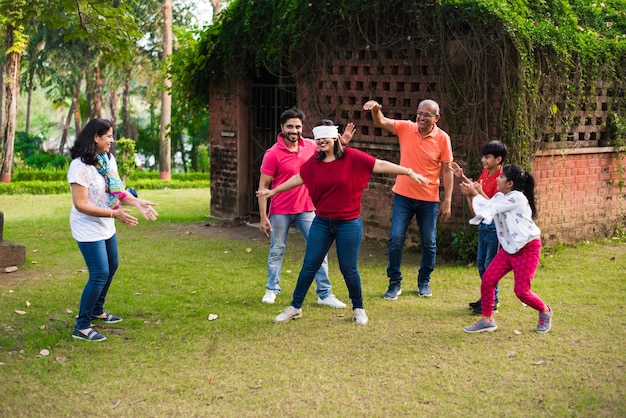 Indische Familie mit verbundenen Augen oder andhali Koshimbir-Spiel im Park oder Garten, asiatische Familie mit mehreren Generationen, die Spaßspiele im Freien spielt