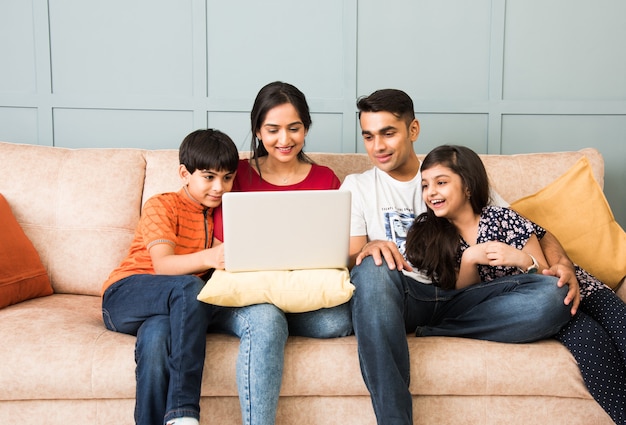 Indische Familie sitzt auf dem Sofa und benutzt Smartphone, Laptop oder Tablet, schaut sich Filme an oder surft im Internet