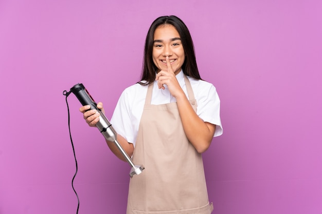 Indische Frau, die Handmixer lokalisiert auf lila Hintergrund tut, der Schweigegeste tut