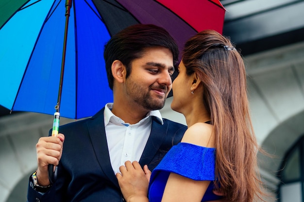 Indische Frau im langen blauen Abendkleid, die romantische Zeit zusammen mit dem hübschen Freund des Liebhabers unter mehrfarbigem Regenbogenregenschirm in der städtischen Innenstadt Europas dreht.