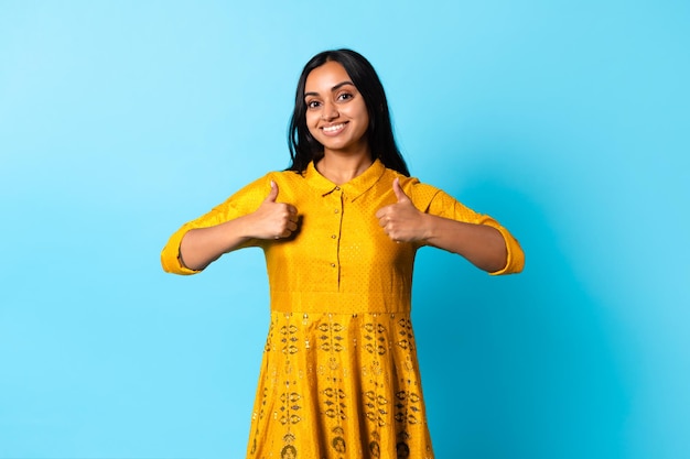 Indische Frau in traditioneller Kleidung, die vor blauem Hintergrund Daumen nach oben zeigt