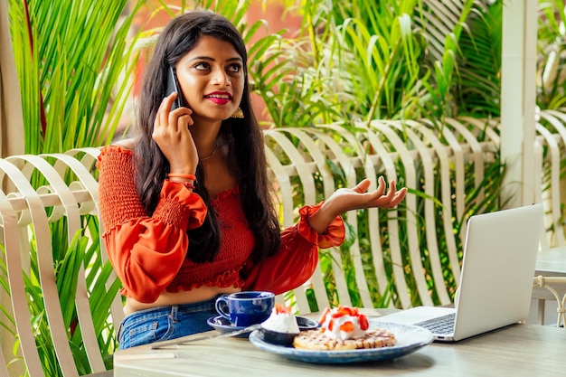 Indische Geschäftsfrau mit Laptop telefoniert im Sommercafé Erfolgsstudent isst Puncakes und trinkt Masala-Tee