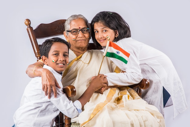 Indische Großmutter und Kinder feiern den Tag der Republik oder den Tag der Unabhängigkeit mit Gesichtern, die mit Trikolore bemalt sind, während sie die Nationalflagge halten, einzeln auf weißem Hintergrund