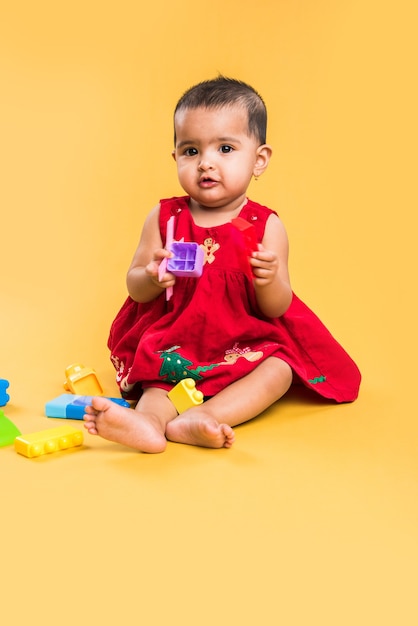 Indische oder asiatische Kleinkinder oder Säuglinge oder Babys, die mit Spielzeug oder Blöcken spielen, während sie liegen oder sitzen, isoliert auf hellem oder buntem Hintergrund