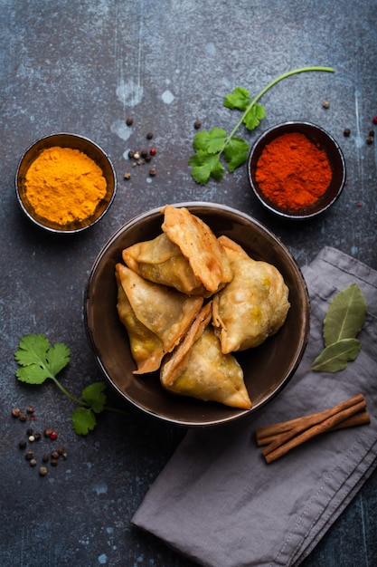 Indische Samosas - gebratenes/gebackenes Gebäck mit herzhafter Füllung, beliebte indische Snacks, serviert in einer Schüssel mit Gewürzen und frischem Koriander auf rustikalem Hintergrund, Draufsicht. Overhead von Samosas