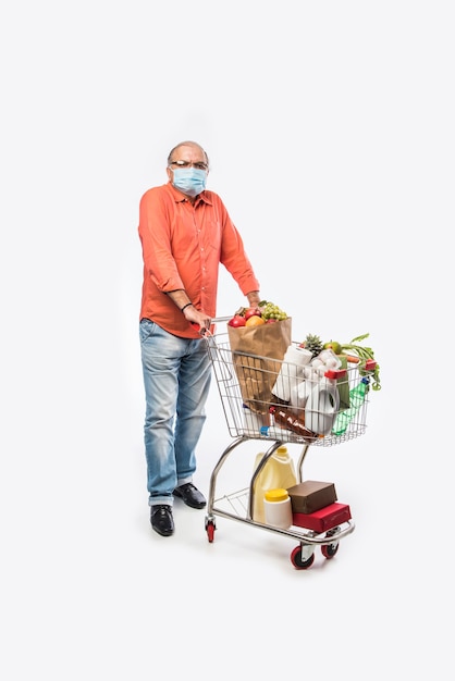 Indischer alter oder älterer Mann trägt eine Gesichtsmaske mit Einkaufswagen oder Trolley voller Lebensmittel, Gemüse und Obst. Isoliertes Foto in voller Länge über weißer Wand