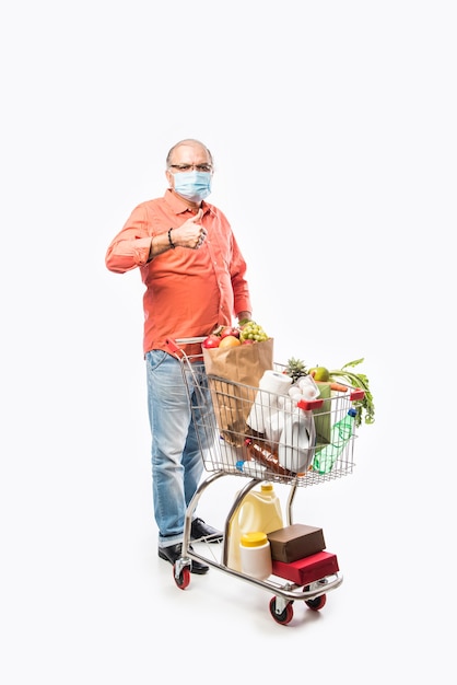 Indischer alter oder älterer Mann trägt eine Gesichtsmaske mit Einkaufswagen oder Trolley voller Lebensmittel, Gemüse und Obst. Isoliertes Foto in voller Länge über weißer Wand