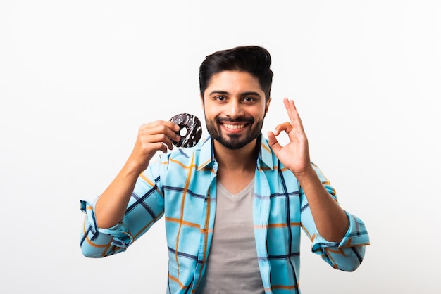 Indischer asiatischer bärtiger junger Mann isst Donut oder Donut, während er am Küchentisch vor gelbem oder weißem Hintergrund sitzt