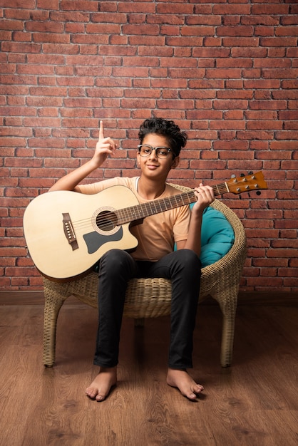 Indischer asiatischer Junge, der akustische Gitarre beim Sitzen gegen weiße Wand spielt