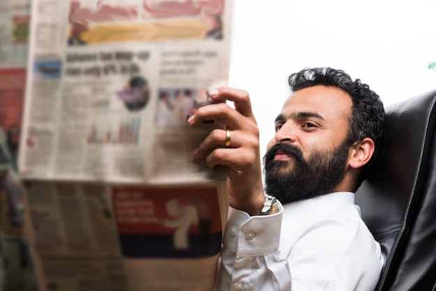 Indischer asiatischer junger Geschäftsmann, der Zeitung liest, während er Kaffee am Schreibtisch trinkt