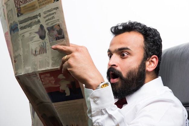 Indischer asiatischer junger Geschäftsmann, der Zeitung liest, während er Kaffee am Schreibtisch trinkt
