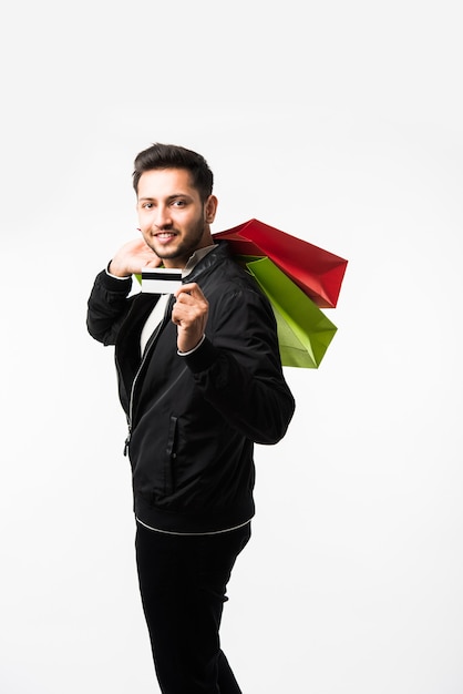 Indischer asiatischer Mann, der seine Einkaufstüten und Kredit- oder Debitkarte zeigt, während er auf weißem Hintergrund steht